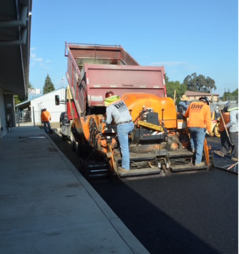 Rear view of a paving crew and machinery laying a continuous mat of fresh black asphalt next to a building