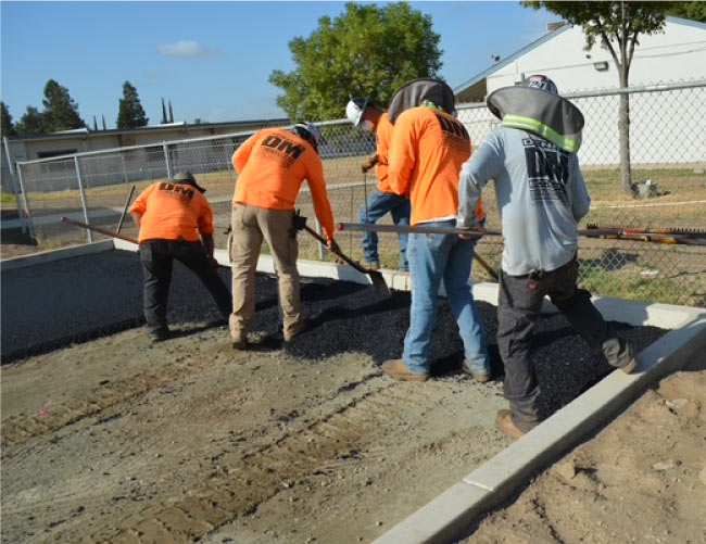Professional paving crew manually grading hot asphalt along a concrete curb to ensure proper drainage and thickness.