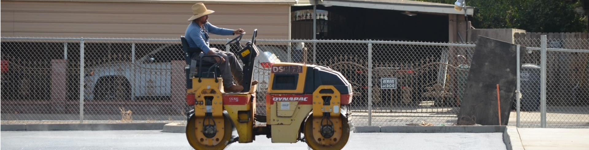 Asphalt paving contractor wearing a straw hat driving a yellow and red Dynapac asphalt roller machine to compact fresh pavement on a job site