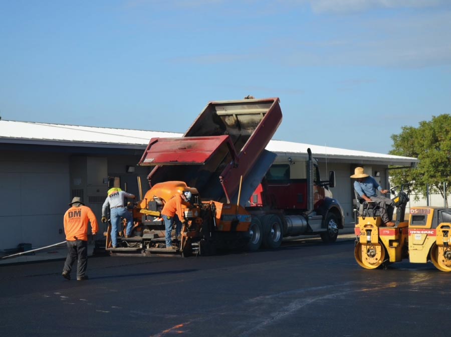 A professional paving crew from Denny McCowan General Engineering Inc., the leading asphalt contractor in selma, ca,  using a red dump truck and an orange paving machine to lay fresh black asphalt on a large commercial lot in the Central Valley.
