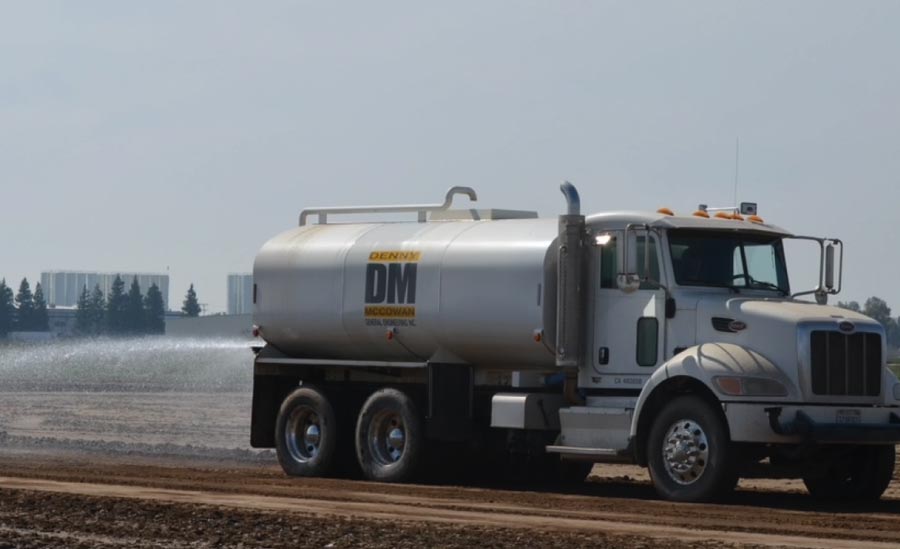 A large white Peterbilt water truck with the "Denny McCowan General Engineering Inc." logo, premier Asphalt Contractor in Clovis, ca. The vehicle is actively spraying water onto a dirt construction site to assist with asphalt grading and dust control. In the background, green trees and several buildings are visible under a clear, hazy sky in the Central Valley.