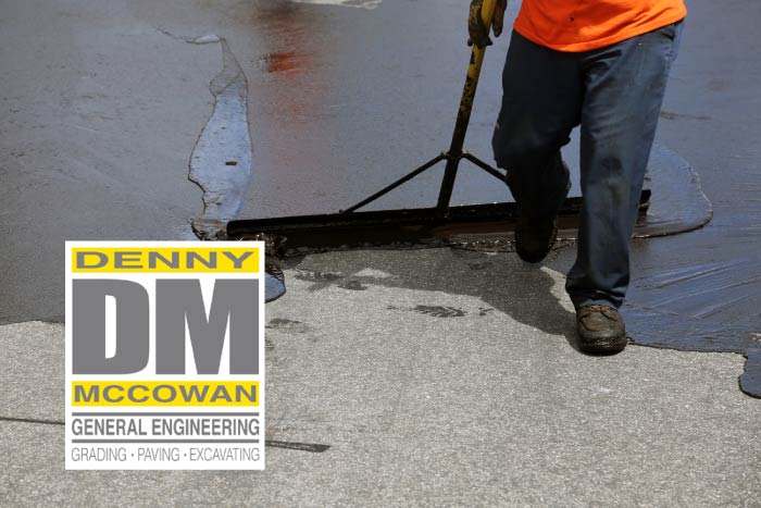Close-up of an experienced crew member meticulously spreading fresh black pavement sealant over an asphalt driveway using an industrial squeegee tool, featuring the Denny McCowan General Engineering logo.