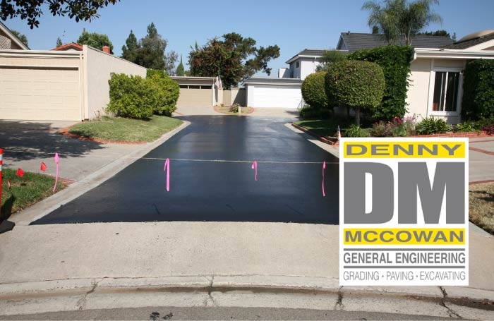 A freshly completed, smooth black asphalt driveway in a residential neighborhood blocked off with caution string for curing, featuring the Denny McCowan General Engineering logo.